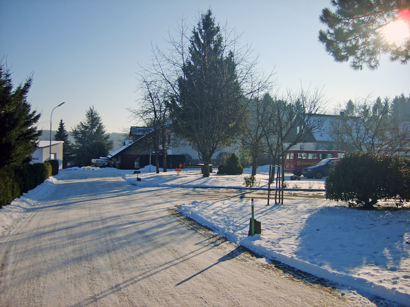 Verschneite Dorfstraße in Stahlhofen am Wiesensee mit Wohnhäusern, Bäumen und blauem Winterhimmel